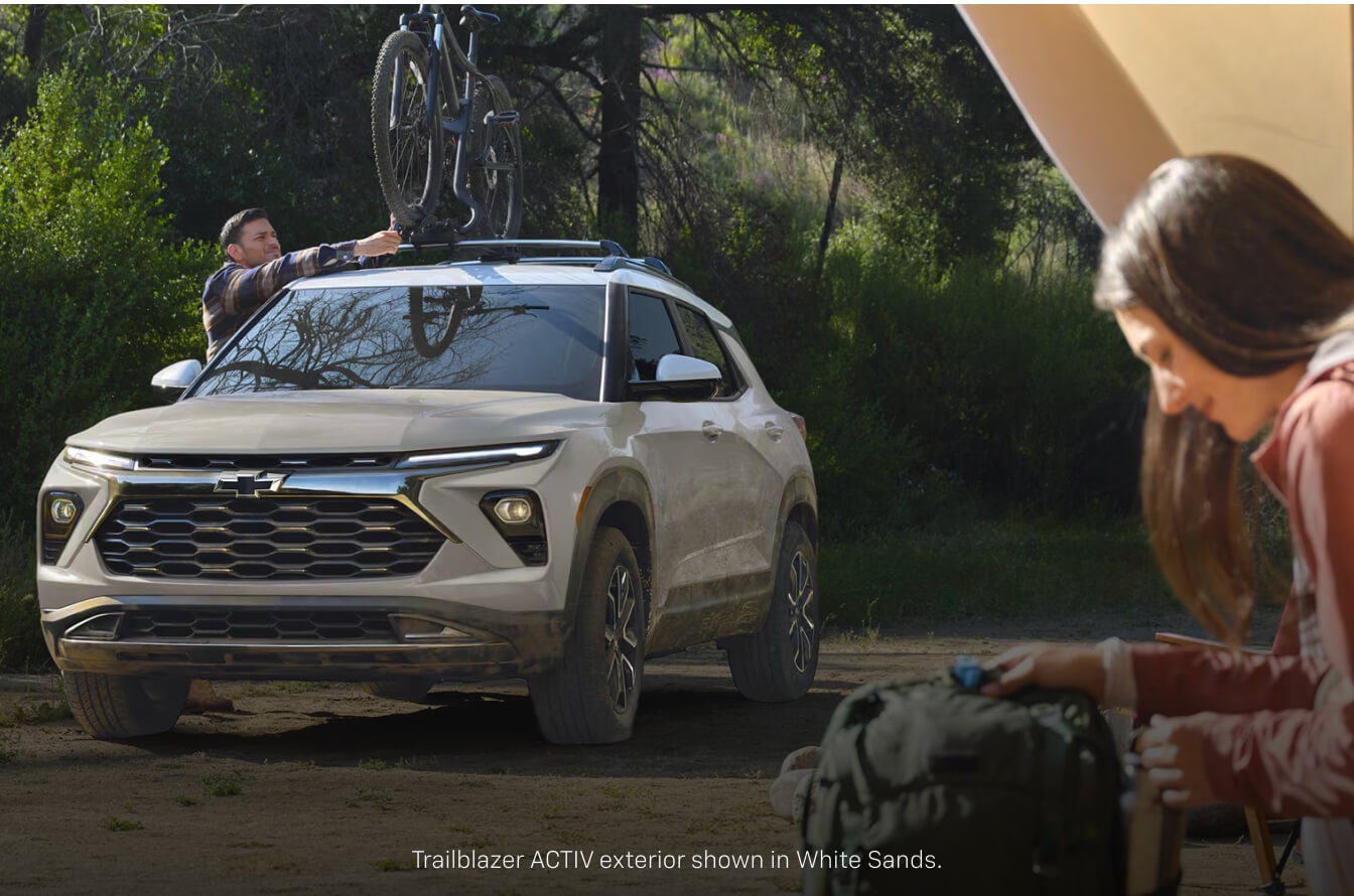 Front view of a sand Chevrolet Trailblazer ACTIV with a roof-mounted bike at a wooded campsite.