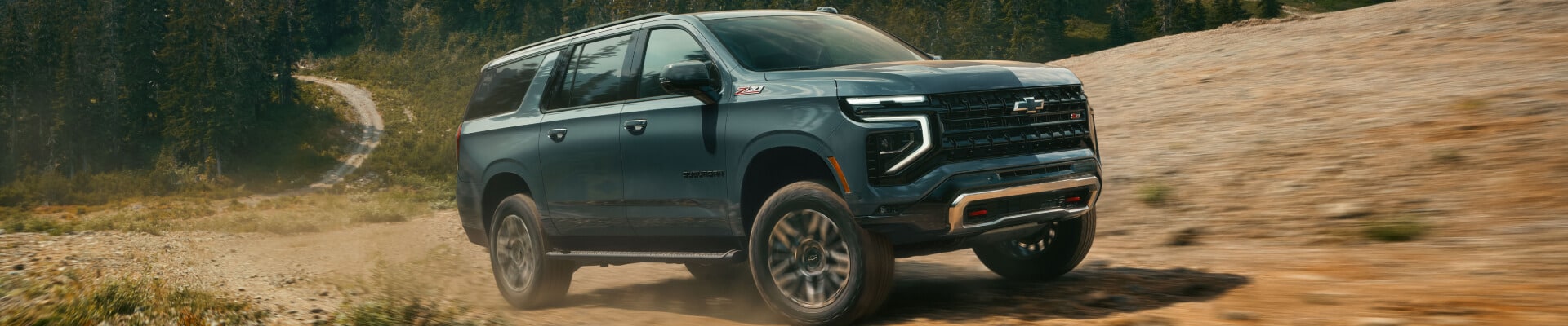 Front three-quarter view of a green 2026 Chevy Suburban Z71 driving on a rugged mountain dirt road.