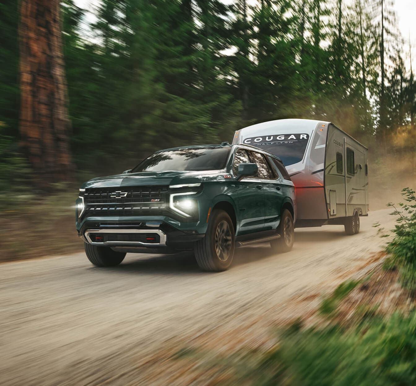 Front three-quarter view of a green 2026 Chevy Tahoe Z71 towing a camper trailer on a dirt forest road.