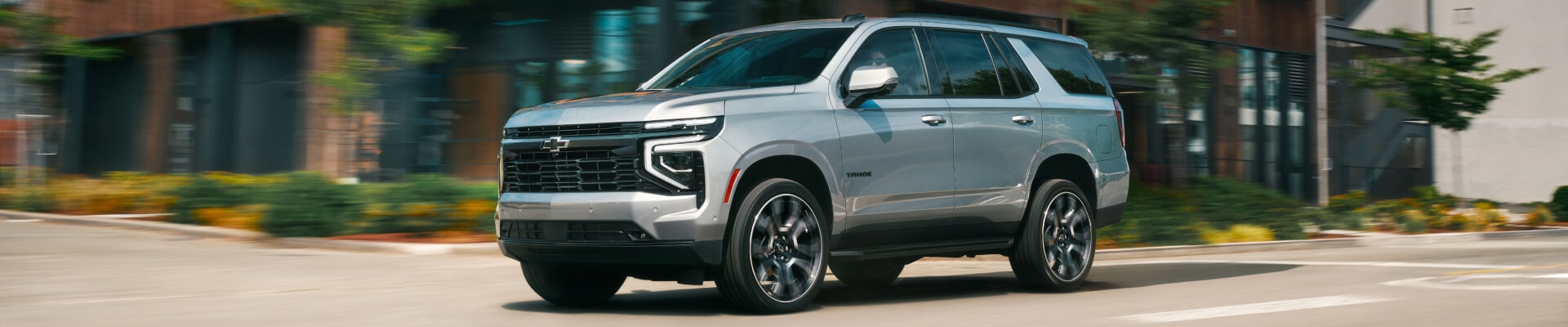 Front three-quarter view of a silver 2026 Chevy Tahoe driving past a modern building.