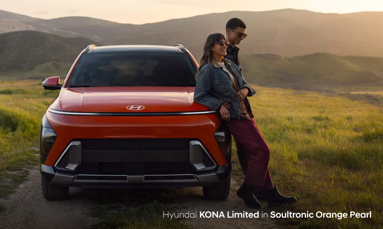 Couple leaning up against their new Hyundai Kona Limited in Soultronic Orange Pearl parked in a grassy plain