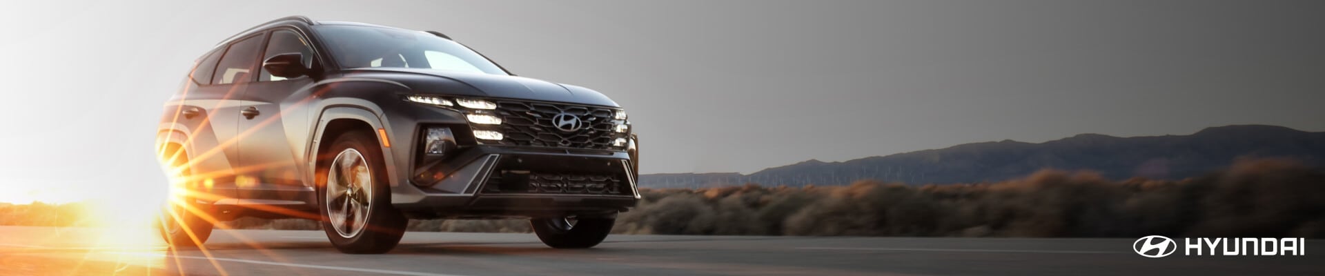 Front three-quarter view of a Hyundai Tucson Limited in Gray driving past mountains at sunset.