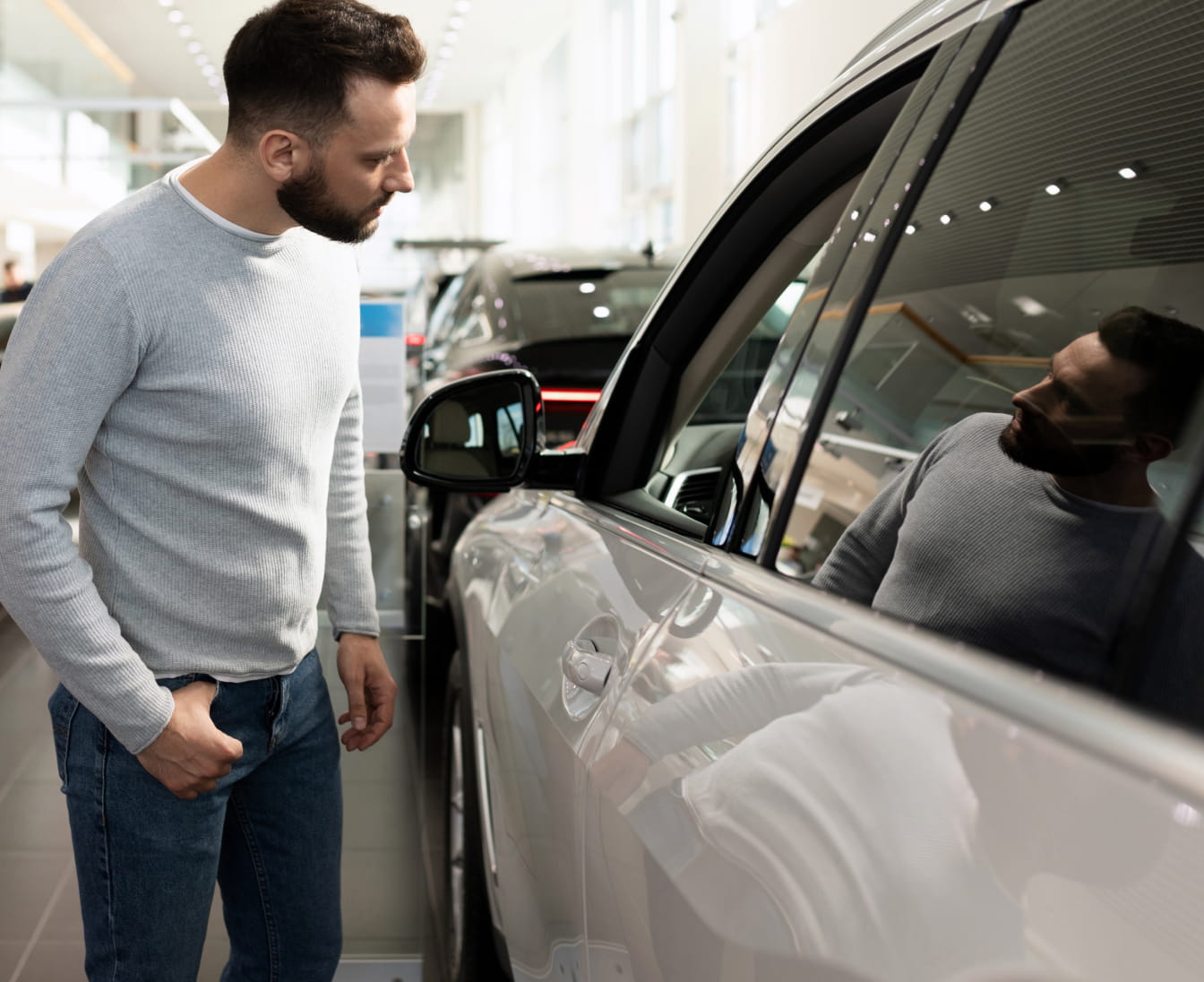 A man in a gray sweater and jeans inspects a shiny car at a dealership. Bright showroom in the background.