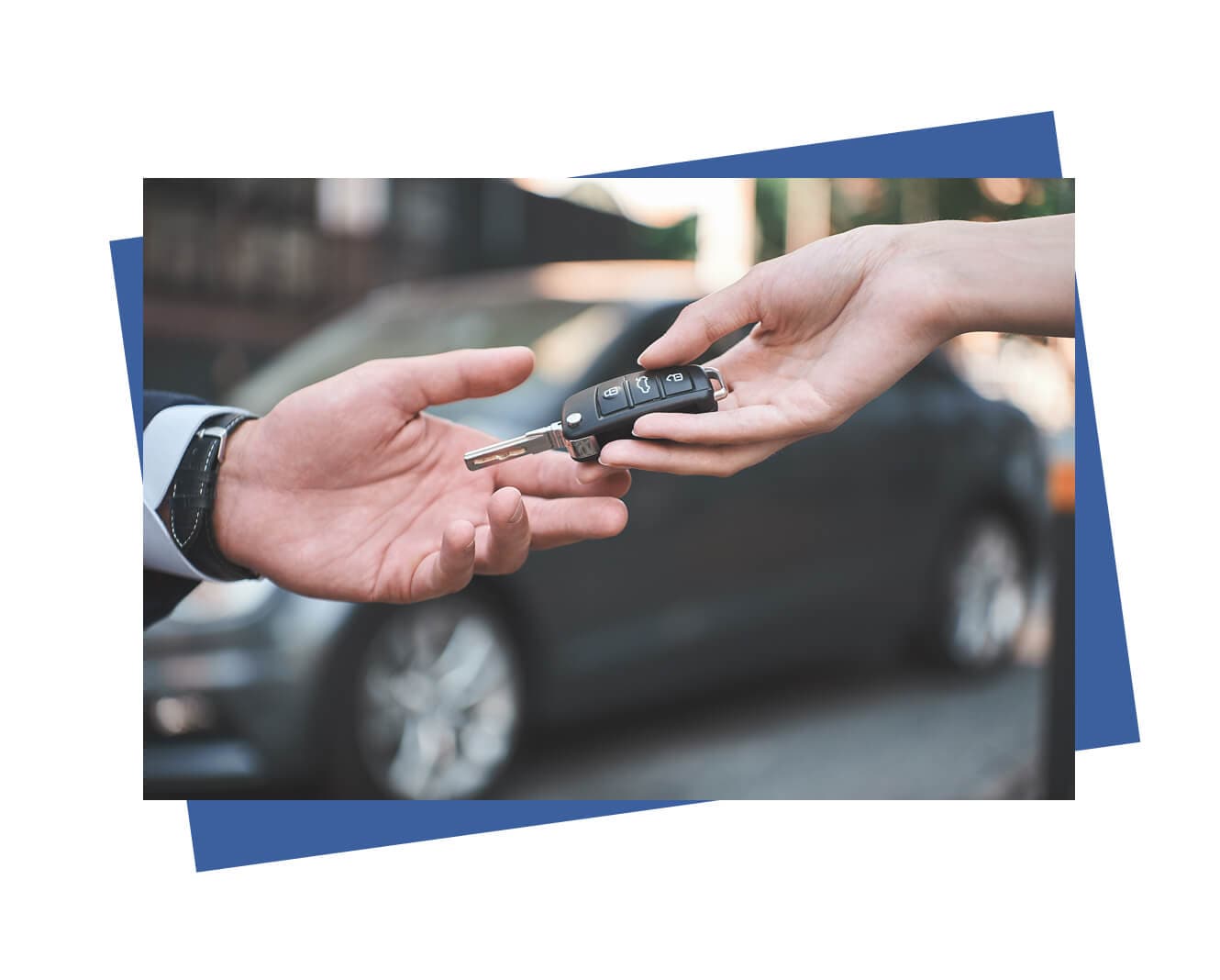A person hands over a car key to another, against a blurred backdrop of a parked car.