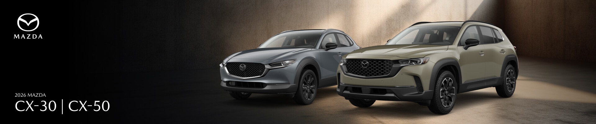 Front three-quarter views of a gray 2026 Mazda CX-30 and a green Mazda CX-50 parked in a dark warehouse.