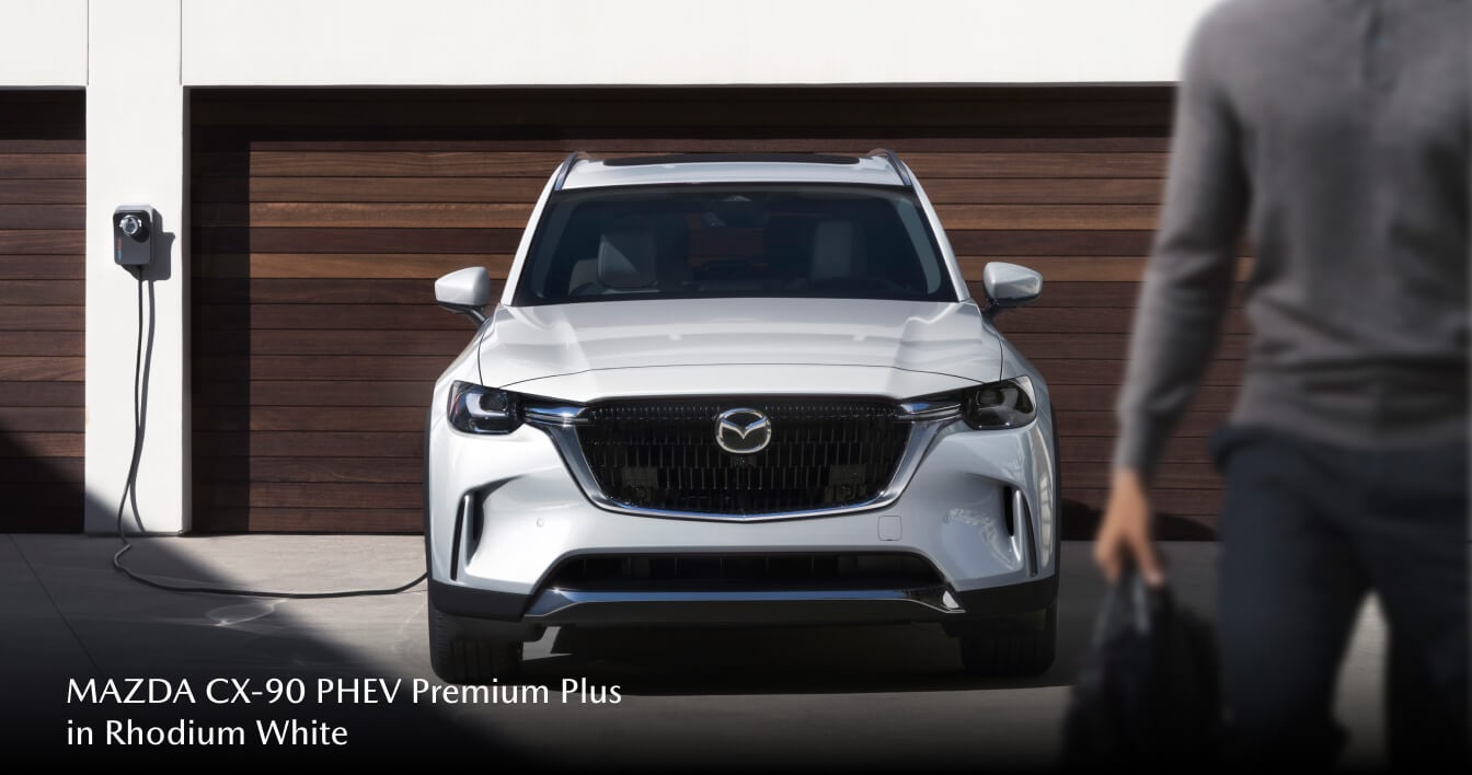 2026 Mazda CX‑90 PHEV Premium Plus in Rhodium White parked and charging in front of a brown garage
