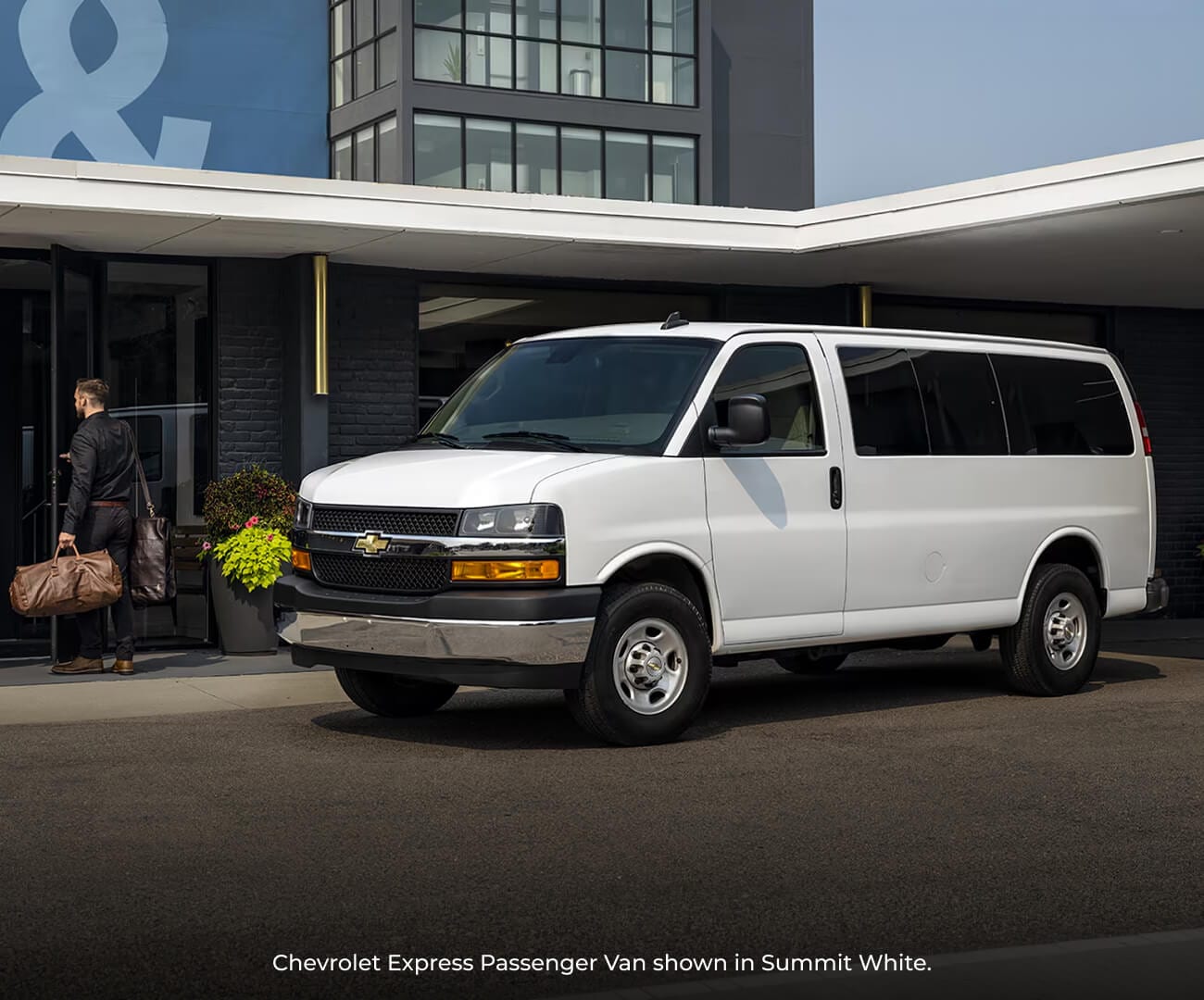 2026 Summit White Chevrolet Express Passenger Van parked at a modern hotel entrance as a man carries a duffel bag inside.