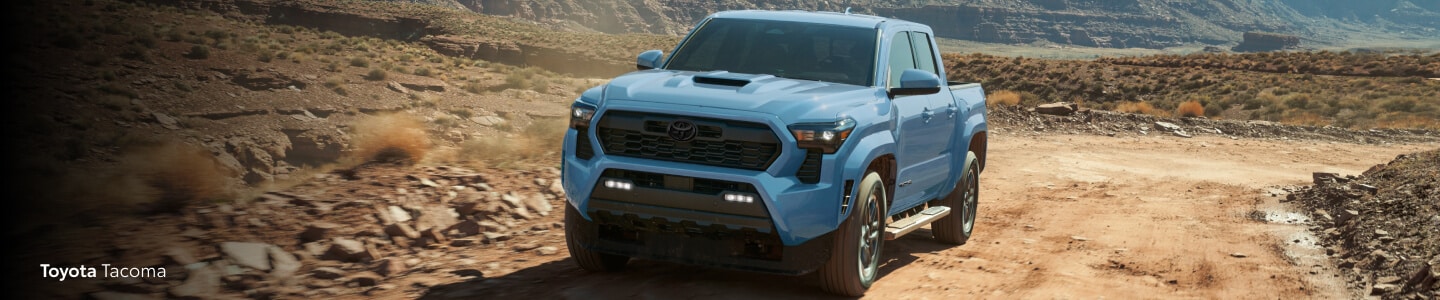 A 2026 Toyota Tacoma TRD Sport in Heritage Blue drives on a rugged, dusty desert road.