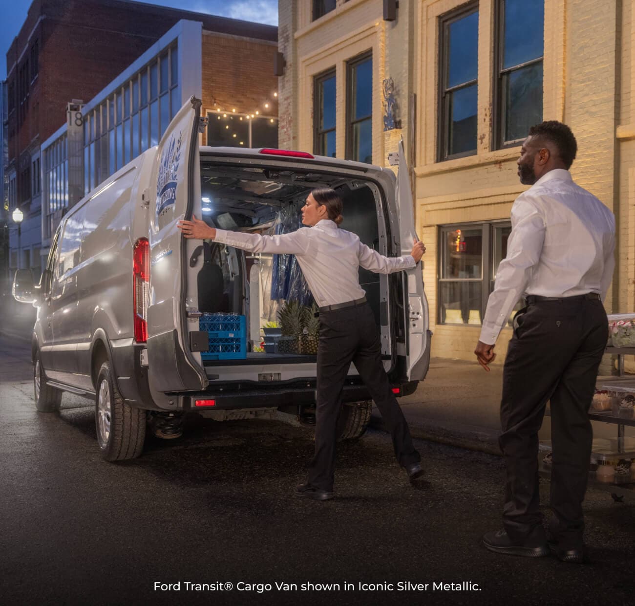 2026 Iconic Silver Metallic Ford Transit Cargo Van parked on a city street as catering workers unload trays.