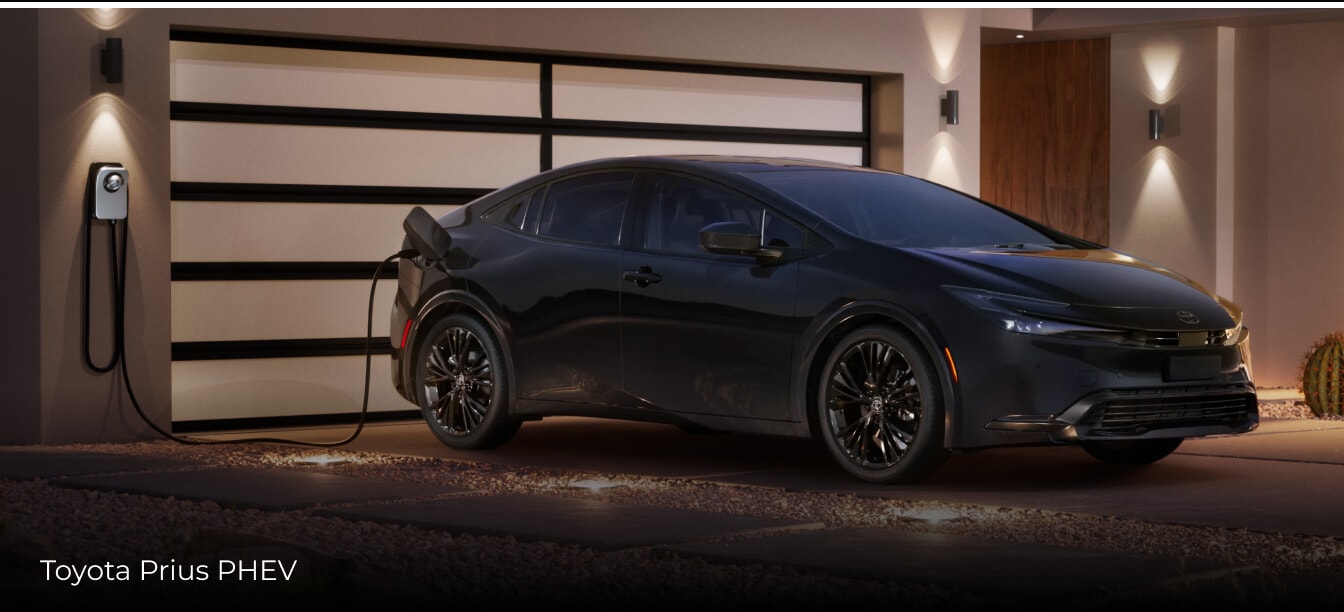 Side view of a black Toyota Prius PHEV plugged in while parked in a driveway.
