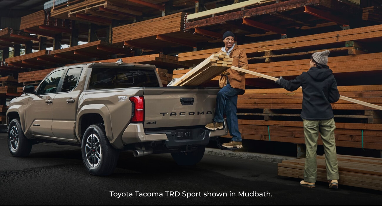 2026 Mudbath Toyota Tacoma TRD Sport truck parked at a lumber yard as two people load wood into the truck bed.