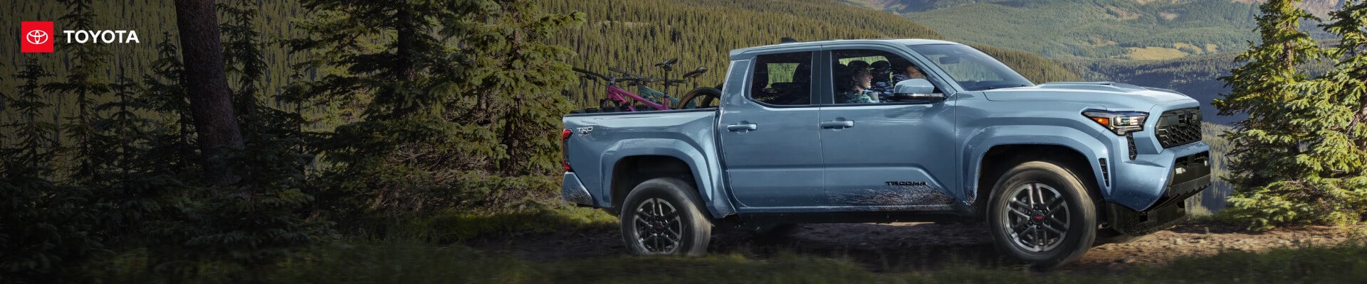 Side profile view of a blue Toyota Tacoma TRD Sport driving over dirt hills in a lush forest.