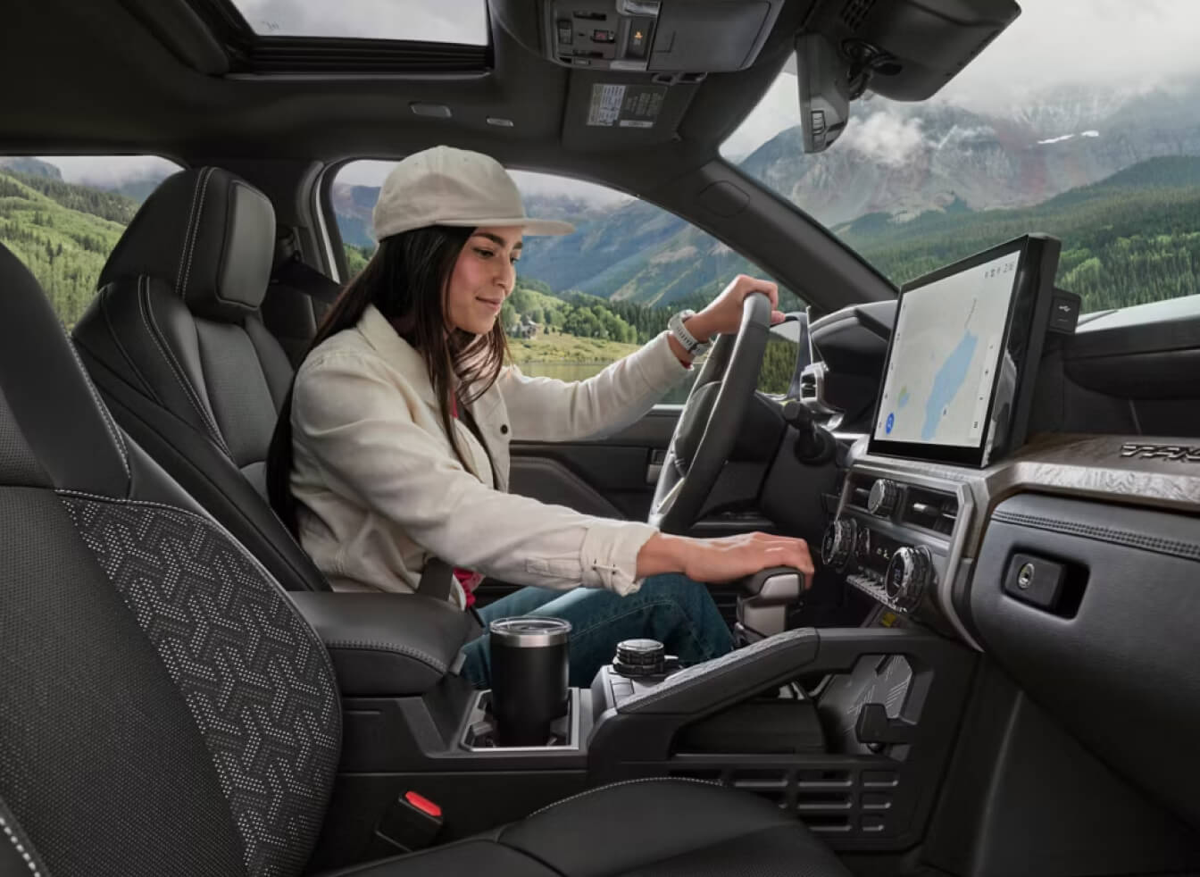 A woman in a cap and jacket sits in the driver's seat of a 2026 Toyota Tacoma Limited in Black SofTex® trim. She is shifting gears. The interior is sleek and modern with a large touchscreen. Outside, scenic mountains and forest are visible.