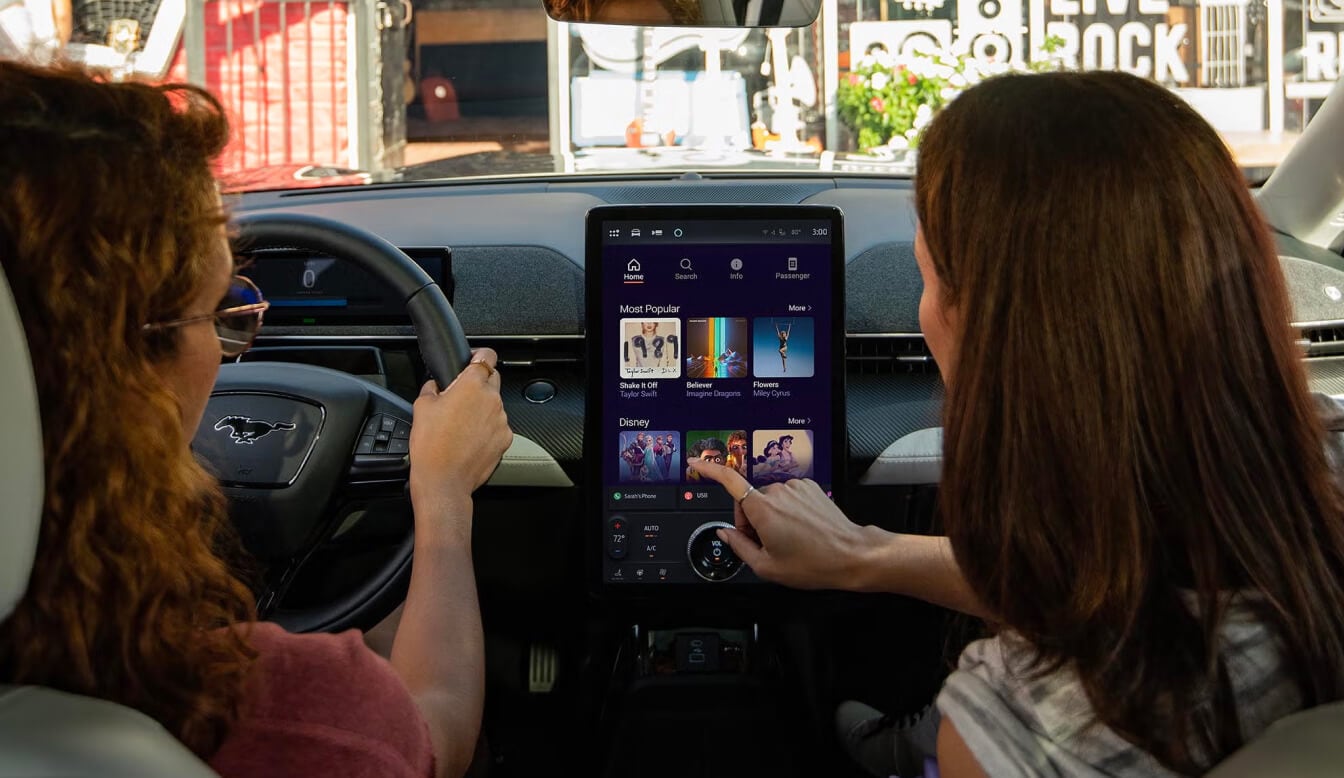 Interior view from back seat of two women interacting with touchscreen infotainment system.