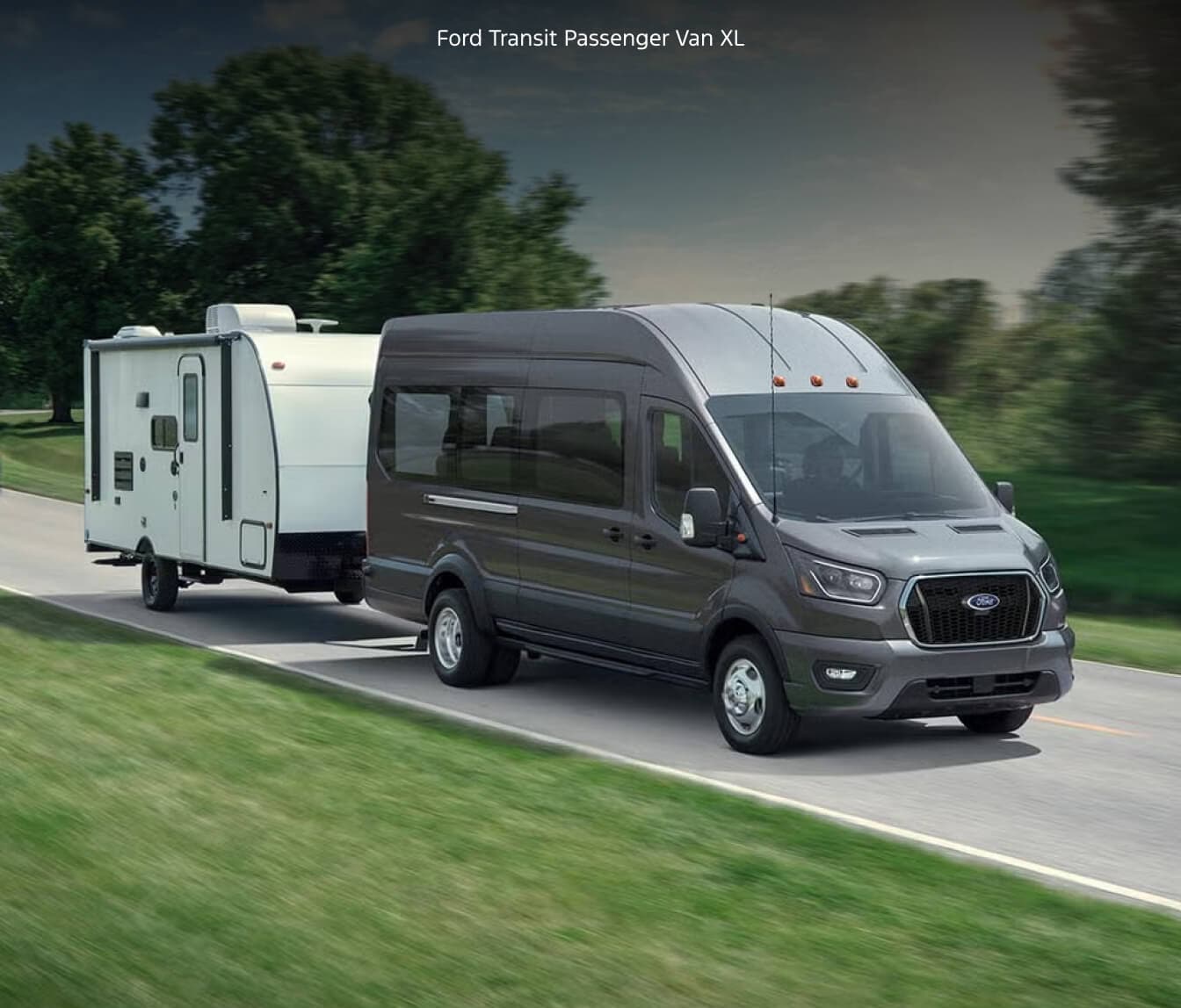 Front three-quarter view of a high-roof 2027 Ford Transit Passenger Van XL in Carbonized Gray Metallic driving along a rural road.