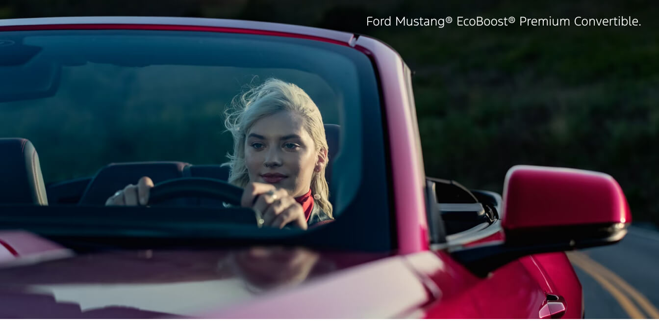 Close-up front view through windshield of a 2026 Red Ford Mustang EcoBoost Premium Convertible with driver behind the wheel.