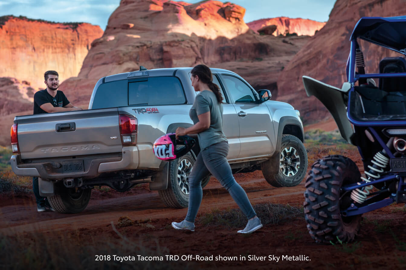 2018 Silver Sky Metallic Toyota Tacoma TRD Off-Road parked in a desert canyon as a woman walks past holding a helmet.