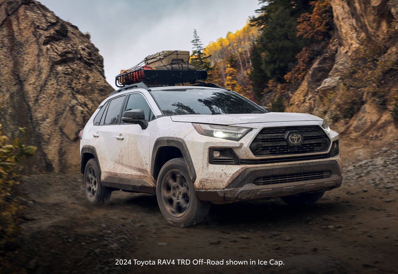 2024 Ice Cap Toyota RAV4 TRD Off-Road driving on a muddy dirt trail between rocky canyon walls.