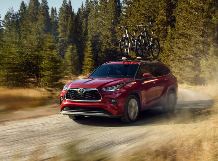 2025 Toyota Highlander Hybrid Platinum in Ruby Flare Pearl driving on a dirt road through pine trees