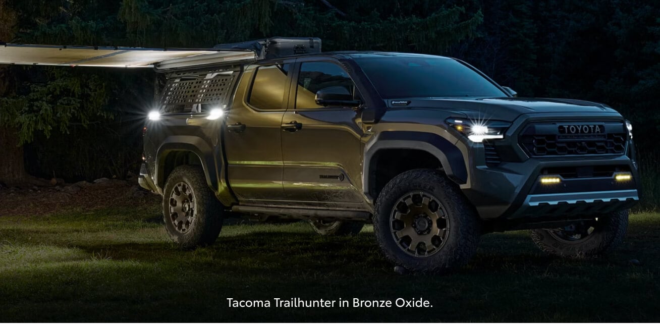 Front three-quarter view of a bronze Toyota Tacoma Trailhunter illuminating a campsite at night.