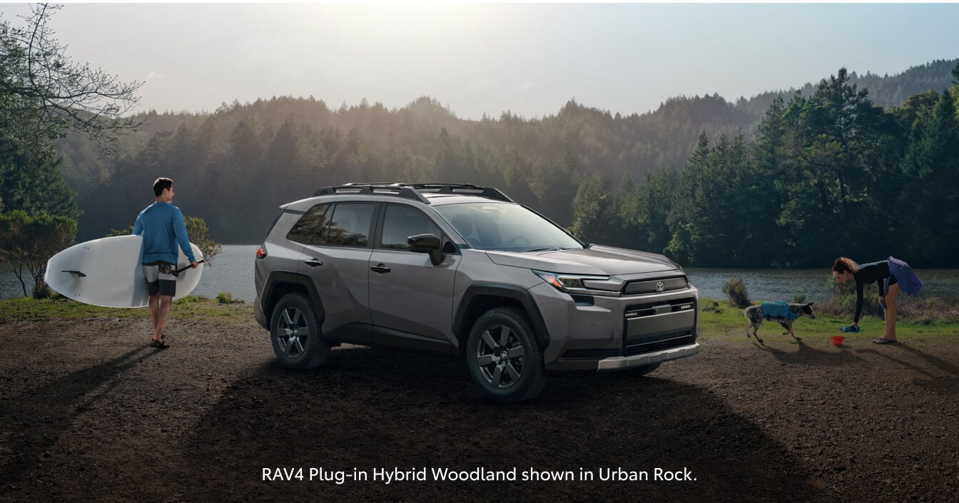 Front three-quarter profile of an Urban Rock Grey Toyota RAV4 Plug-In Hybrid Woodland Edition parked on a lakeside shore.