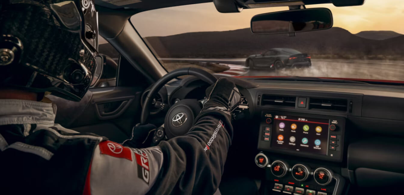 An interior shot over the shoulder of a driver in track safety gear driving a Toyota GR86 on a smokey racetrack.