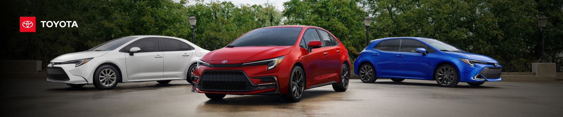 A white 2023 Toyota Camry, red Corolla and blue Corolla hatchback parked in a tree-lined lot.