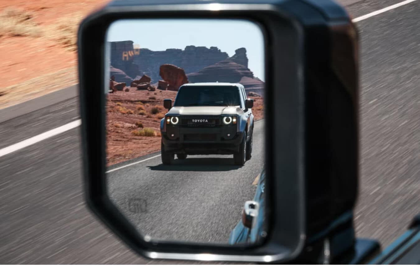 Side-view mirror reflection of a Toyota Land Cruiser 1958 in Trail Dust driving along a paved road.