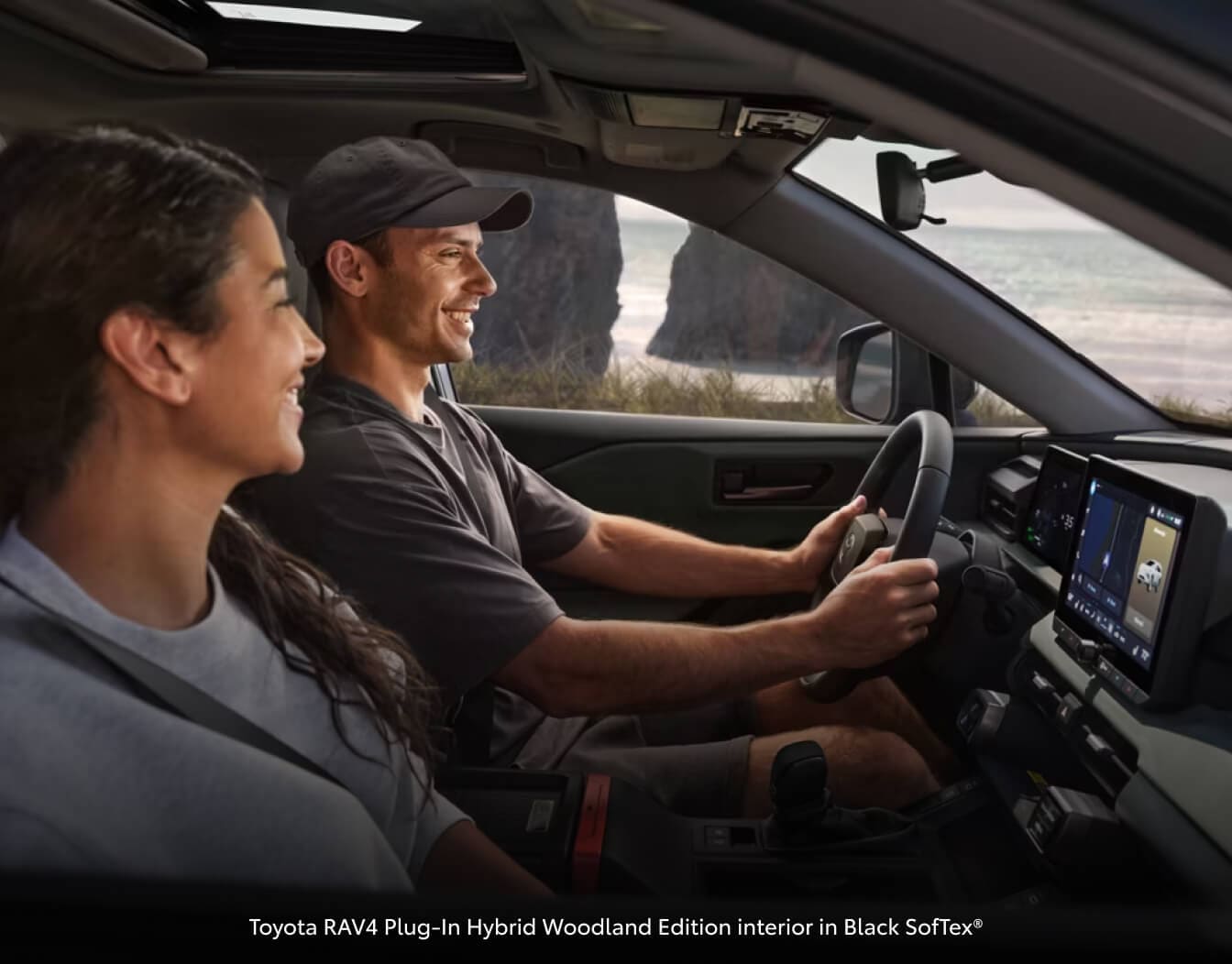 Toyota RAV4 Plug-In Hybrid Woodland Edition interior in Black SofTex® showing a side view displaying the dashboard touchscreen near the ocean.