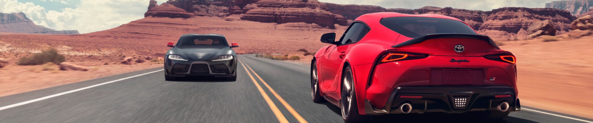 Rear and front views of a red 2026 Toyota GR Supra and black GR Supra GT4 driving on a desert canyon road.