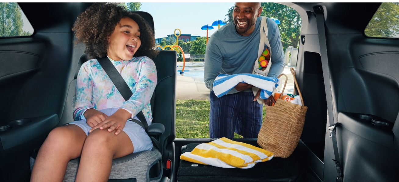 A toddler with her seatbelt on in the back of a vehicle with her father unloading the truck behind her