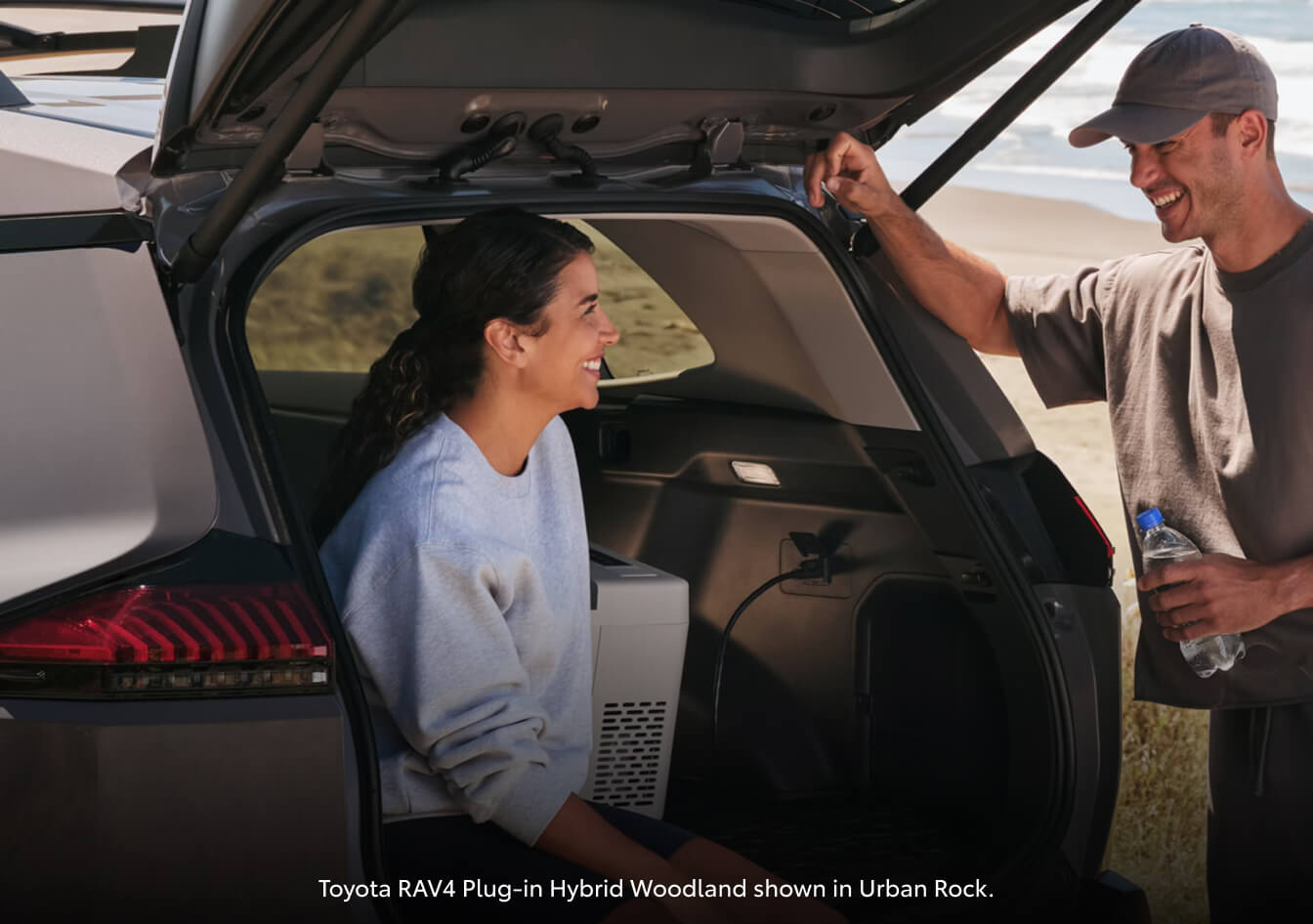 Close-up of a happy couple by the open cargo liftgate of an Urban Rock grey Toyota RAV4 Plug-in Hybrid Woodland powering a portable cooler.