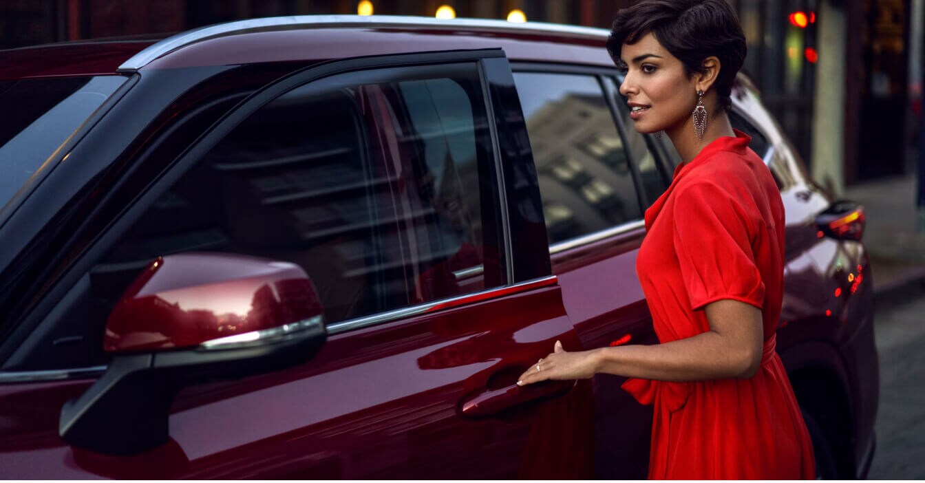 Side view of a 26 Toyota Highlander Platinum in Ruby Flare Pearl as a woman opens the door on a city street.