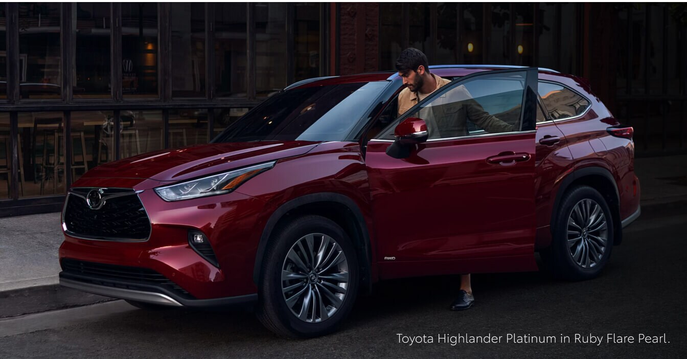 Front quarter view of a 26 Toyota Highlander Platinum in Ruby Flare Pearl as a driver exits the vehicle on a city street.