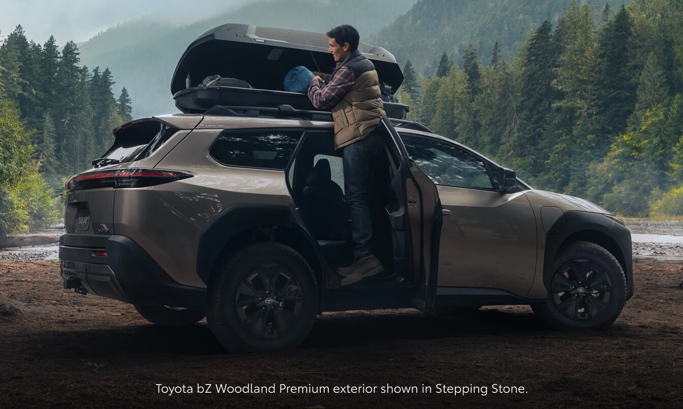 Rear three-quarter view of a Stepping Stone 2026 Toyota bZ Woodland Premium as a man packs a roof cargo box.