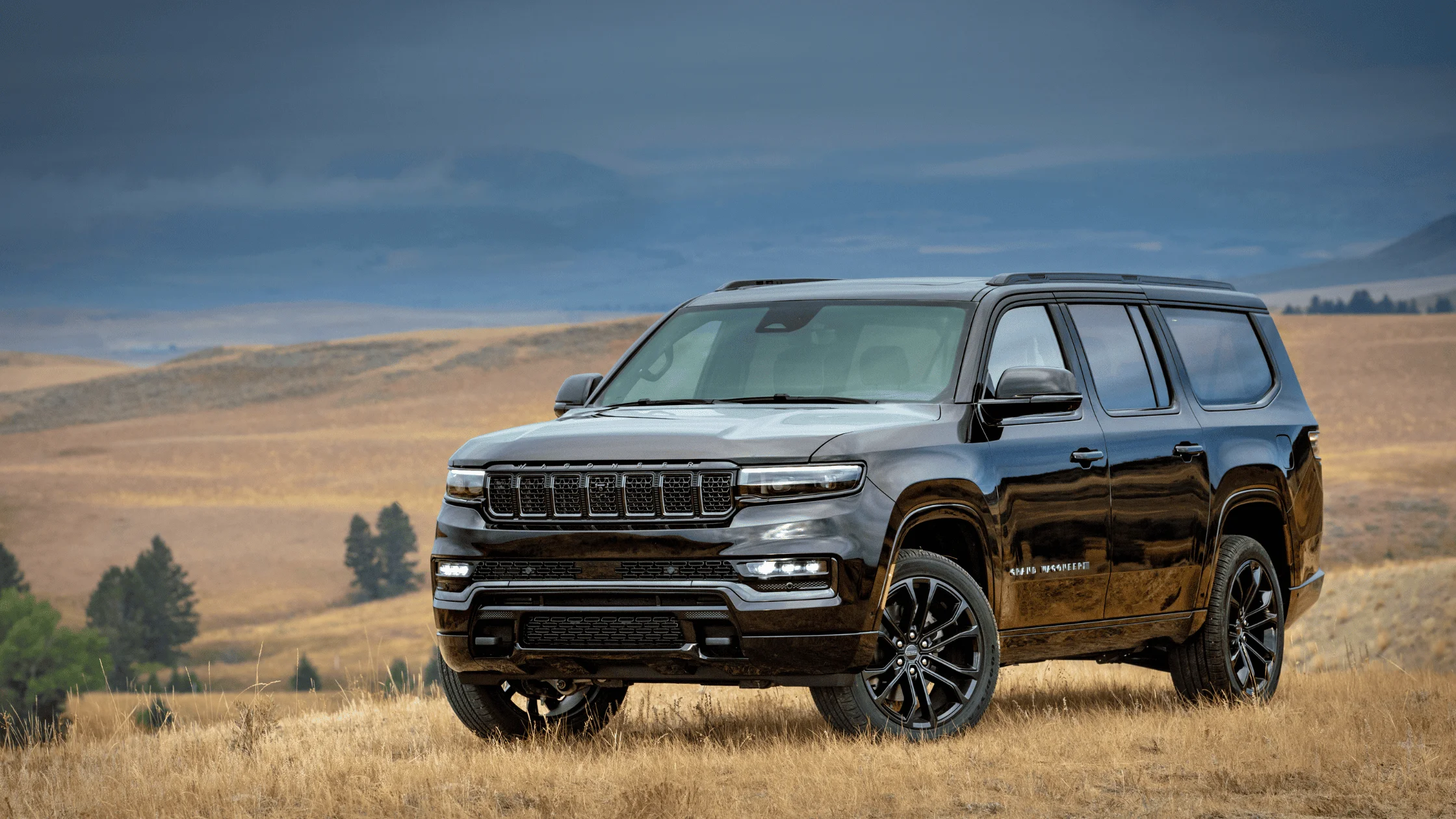 lack 2026 Jeep Grand Wagoneer SUV parked on dry grassland, highlighting its luxury design and bold seven-slot grille under a dramatic sky.