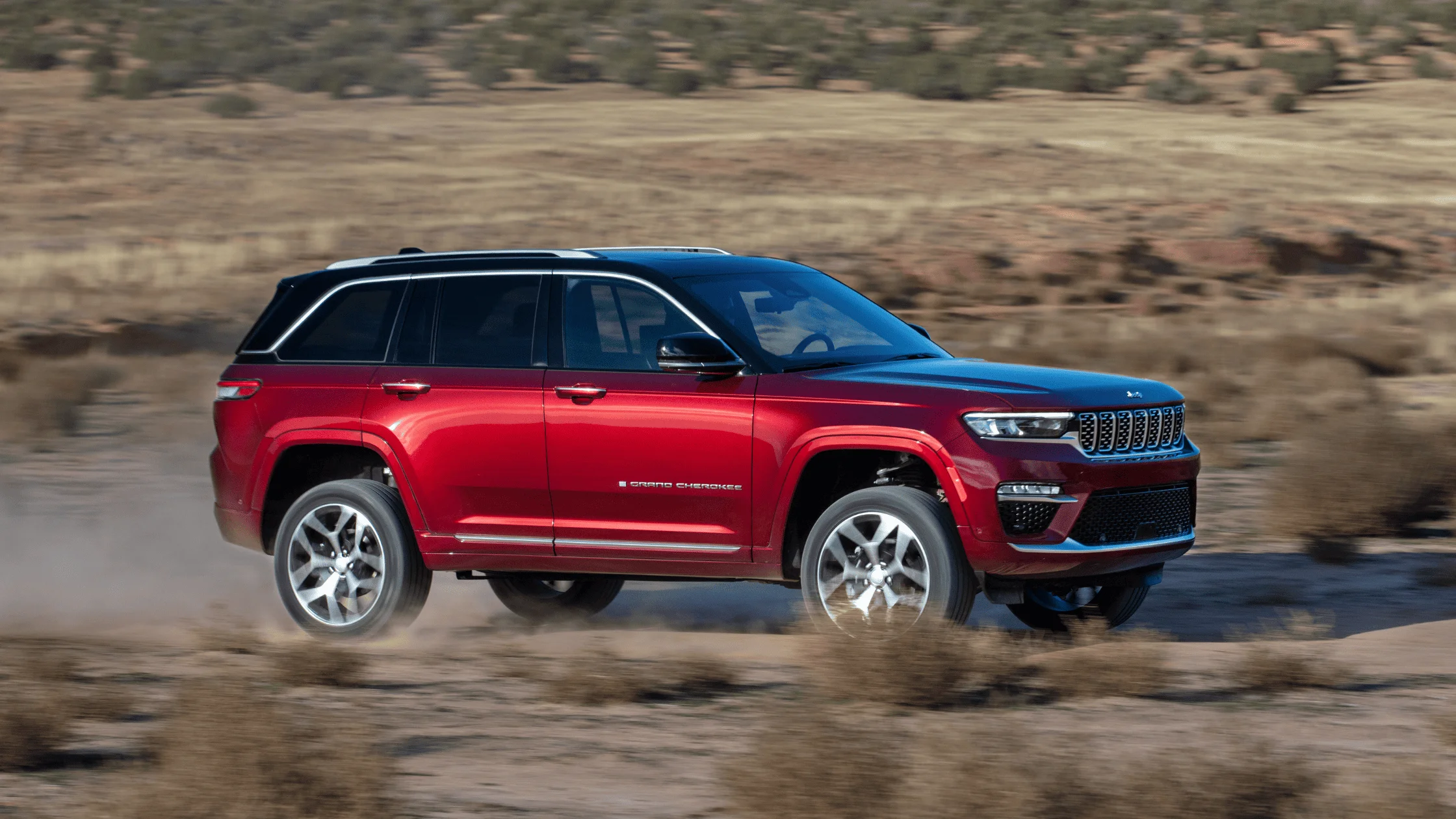 2026 Jeep Grand Cherokee in red driving through dry brush landscape with motion blur on dirt trail