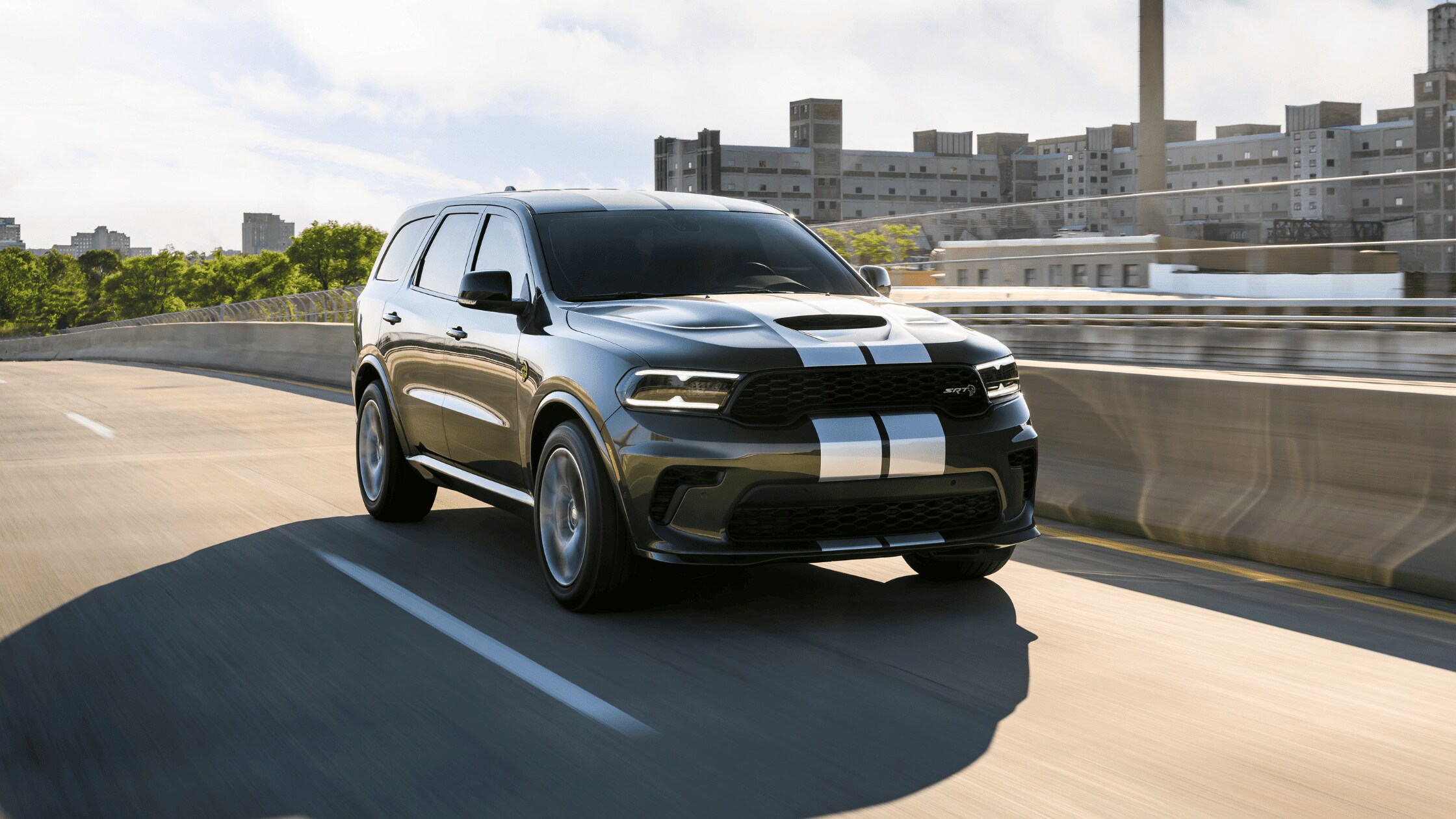 Black Dodge Durango SUV with white stripes on highway, front angle city backdrop