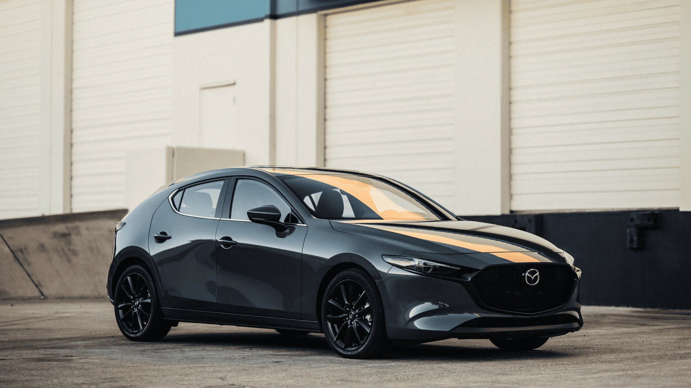 The 2025 Mazda3 Hatchback in Machine Gray Metallic parked near an industrial building, emphasizing its aerodynamic lines, gloss-black wheels, and compact performance design.