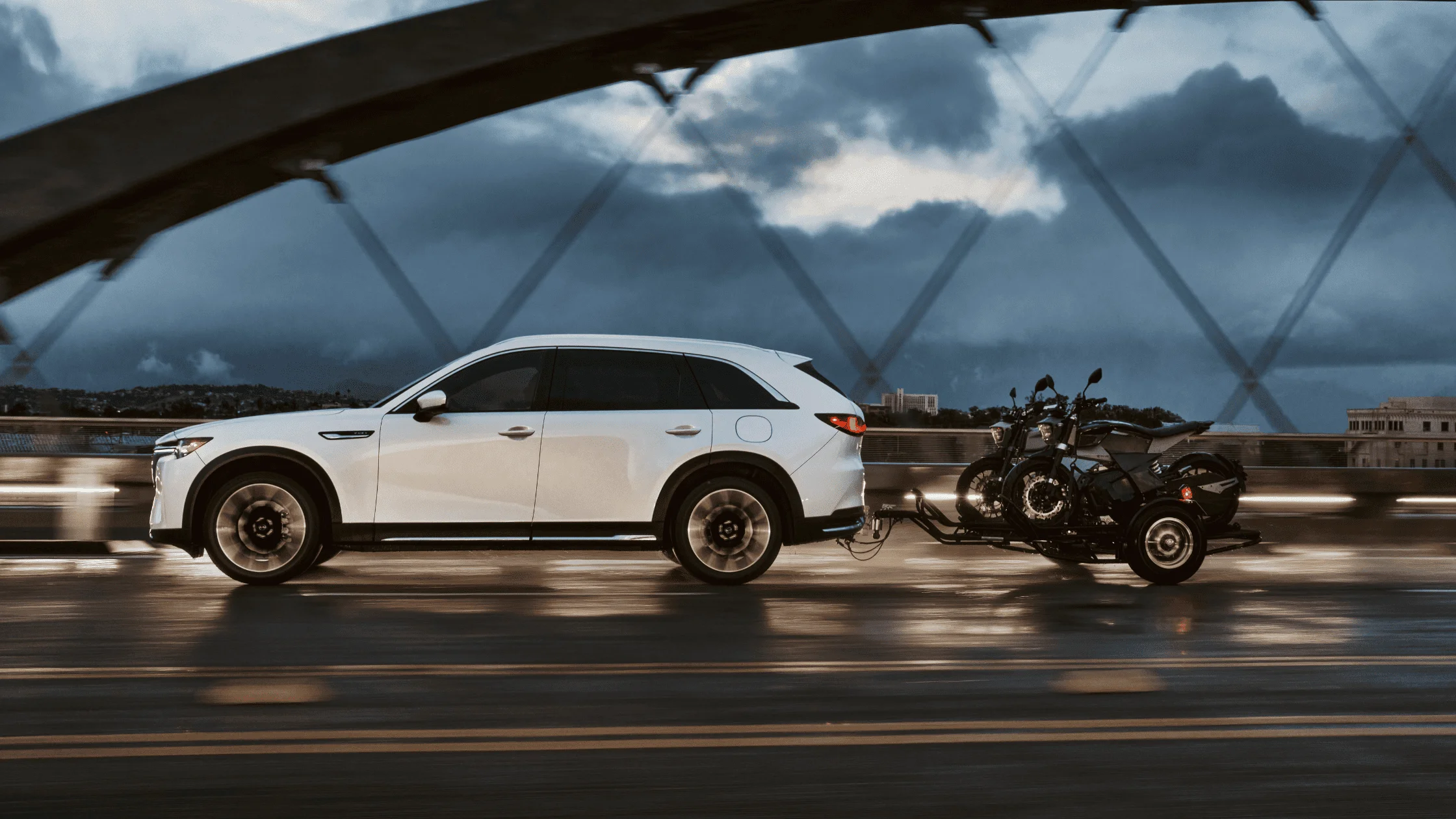 The 2025 Mazda CX-90 in white tows two motorcycles across a modern bridge at dusk, highlighting its premium styling, powerful stance, and impressive towing capability.