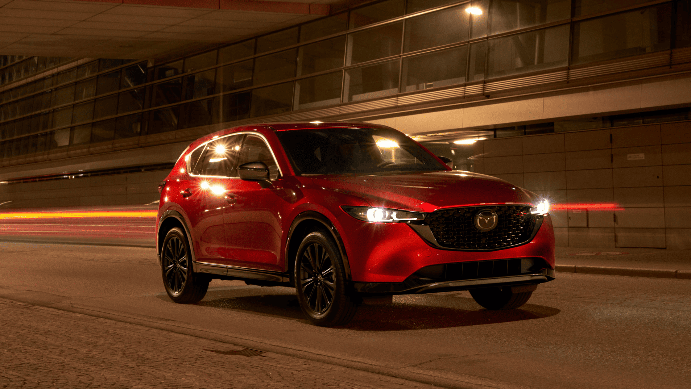 Red Mazda CX-5 SUV at night, exterior front view with headlights on under city lights