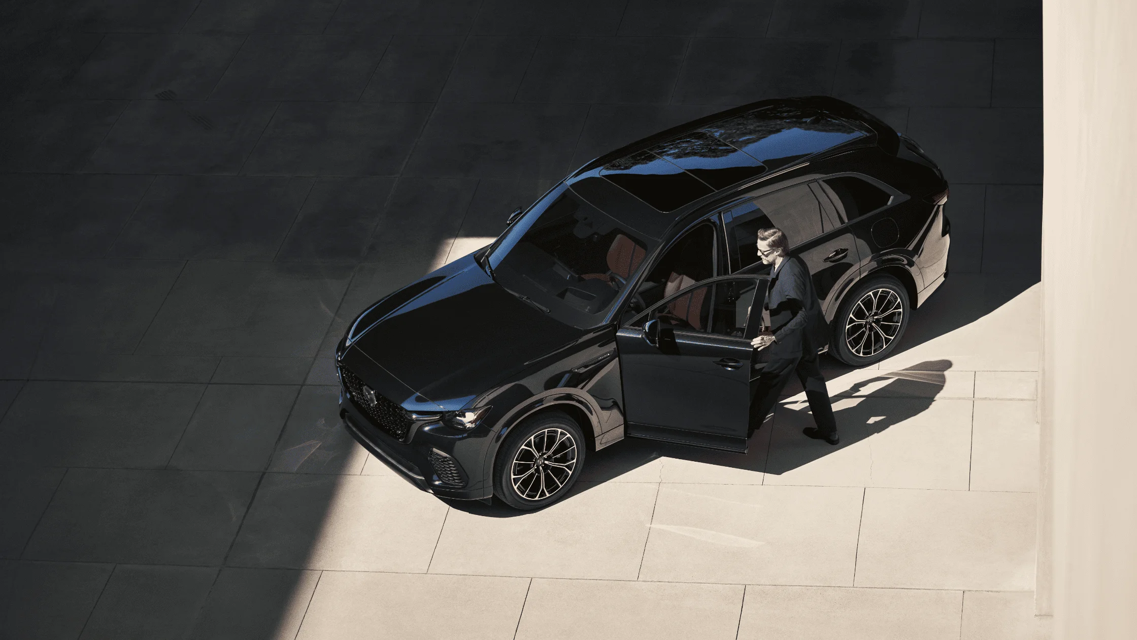 Overhead view of 2025 Mazda CX-70 in black with a man opening the driver door, parked in shadowed concrete plaza