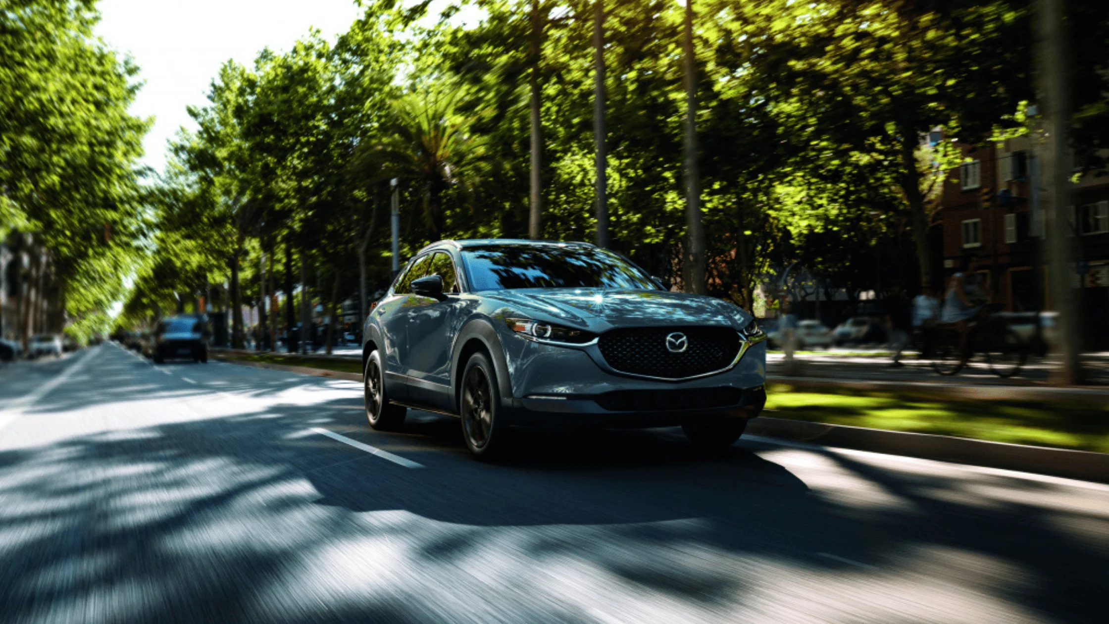 2025 Mazda CX-30 in gray driving through sunlit urban boulevard lined with trees and blurred motion background