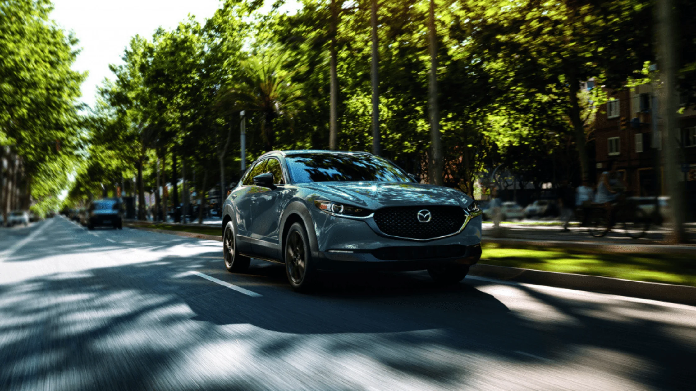 The 2025 Mazda CX-30 in Polymetal Gray drives along a sunlit city boulevard lined with trees, showcasing its sleek design, LED headlights, and refined front fascia in motion.