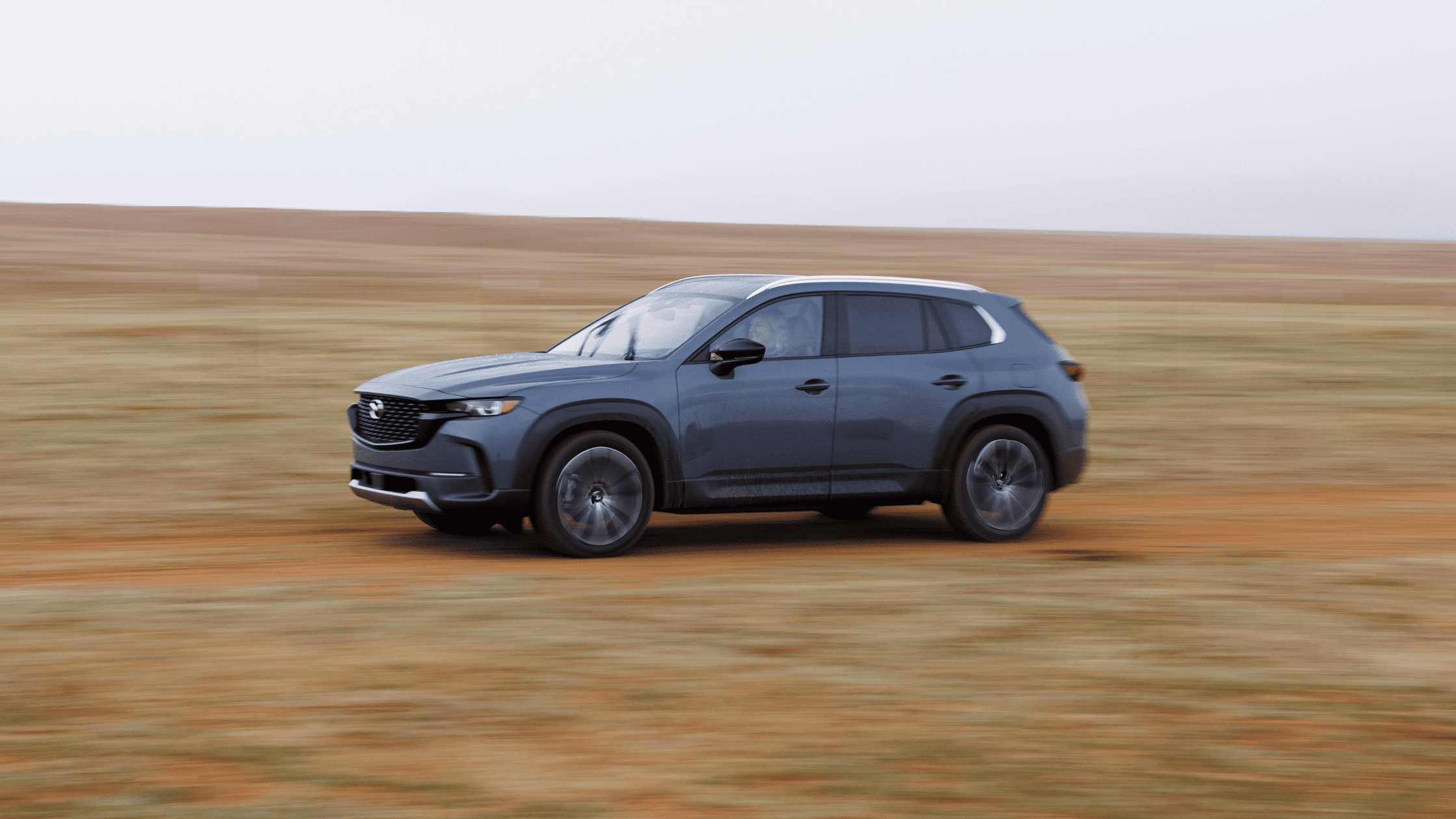 Blue Mazda CX-50 SUV driving on dirt trail, side profile motion shot in open terrain