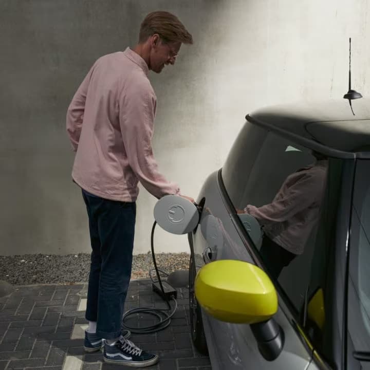 Man in pink button-down shirt and Vans shoes standing on a brick surface and plugging a charging cord into a MINI Electric vehicle, with a shadowed white wall in the background.