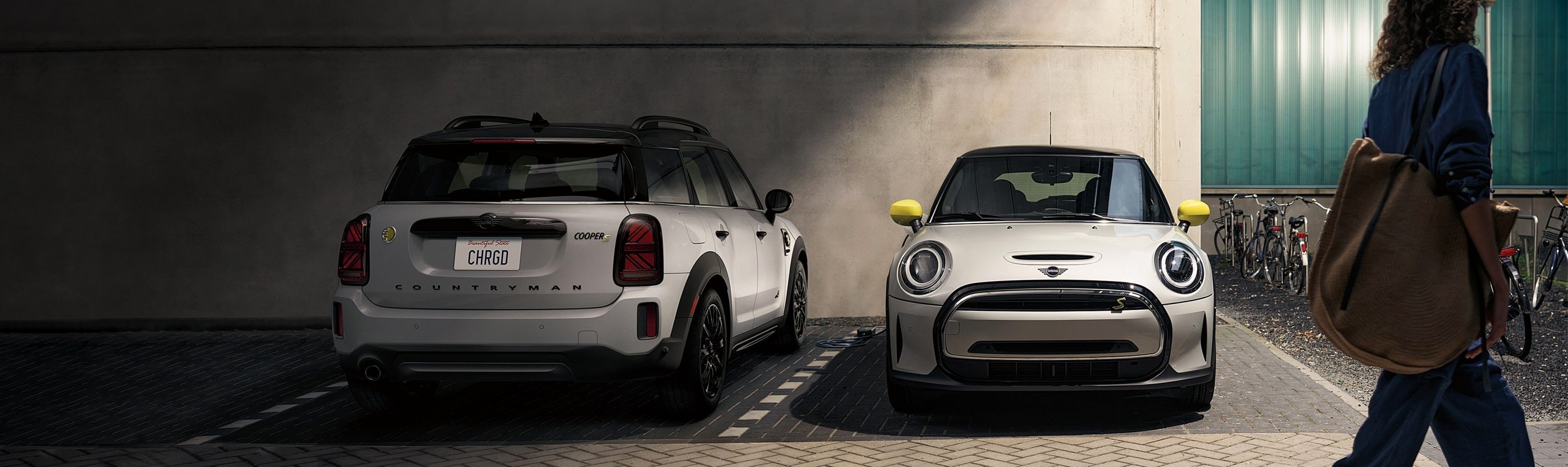 Two MINI vehicles, one back view of a MINI Countryman with an overcast shadow and one front view of a MINI Countryman, parked in designated parking spots on a cobblestone surface outside a building with a woman walking past on the right side.