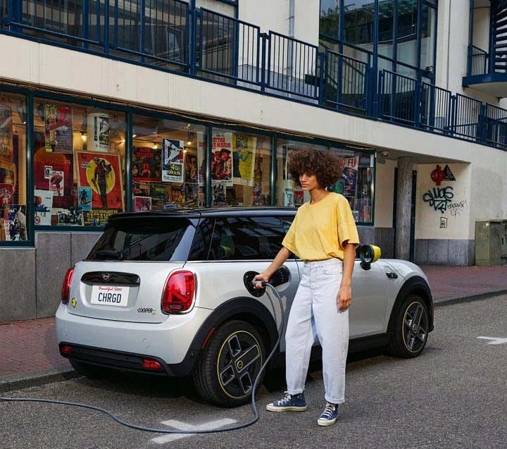 Three-quarters back view of a white MINI Cooper SE Hardtop 2 Door vehicle parked on a street, with a person in a yellow shirt plugging a charger into the vehicle plus a windowed retail storefront in the background.
