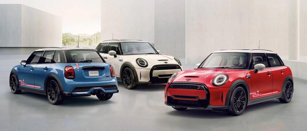 Three MINI 20 Years Edition cars in blue, white, and red, parked side by side on a grey surface with white square buildings in the background.