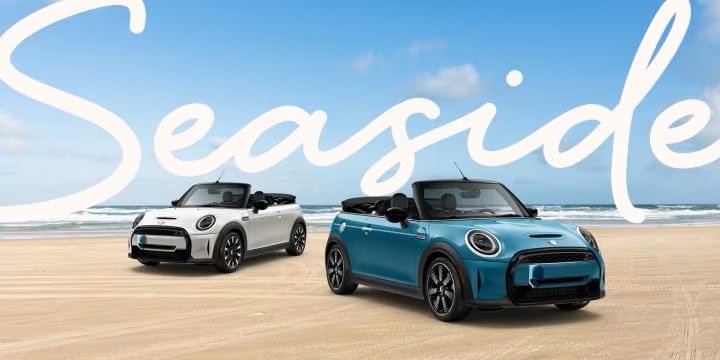 Two MINI Seaside Edition vehicles side by side, the left one (Nanuq White) in a three-quarter front-left view and the right one (Caribbean Aqua) in a three-quarter front-right view, parked on a sandy beach.